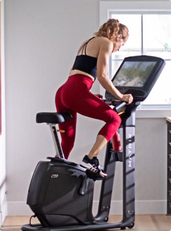 
                        
                         Woman exercising on a black exercise bike in a home setting.
                        
                        